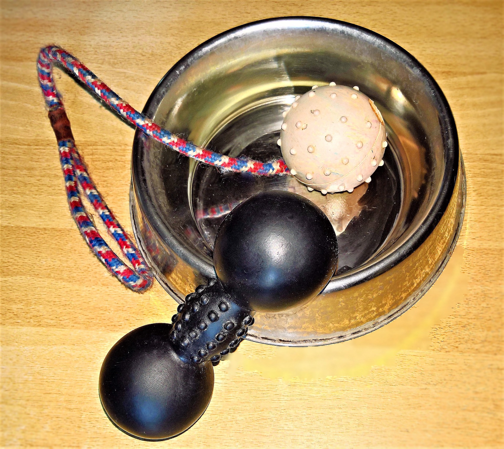 A dog bowl on a wooden surface full of dog toys: a rubber black toy with both ends rounded and the connective middle cylinder covered in raised bumps and a round tan ball also with raised bumps connected to a red, white and blue striped rope with a loop on one end