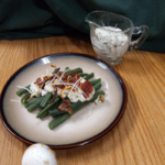 A tan plate with green beans topped with white sauce, bacon, cheese; a small crystal gravy dish with white sauce in the back right and a white mushroom in the front with a deep green background and wood table