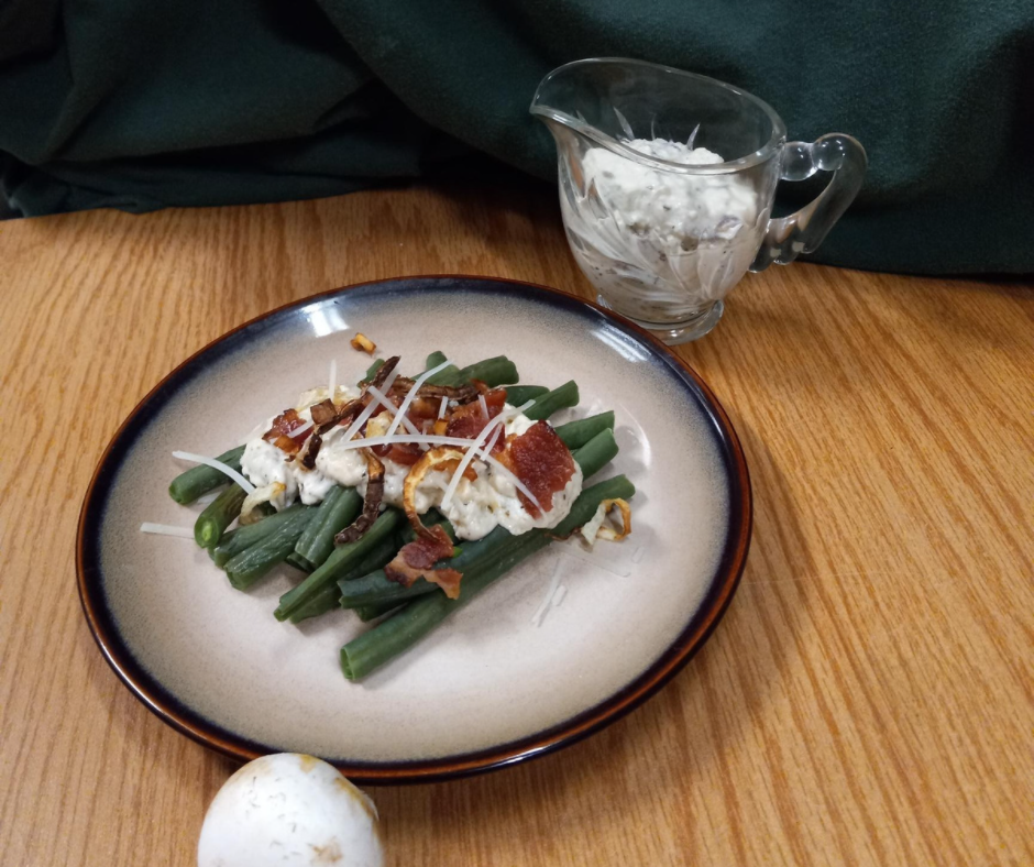 A tan plate with green beans topped with white sauce, bacon, cheese; a small crystal gravy dish with white sauce in the back right and a white mushroom in the front with a deep green background and wood table