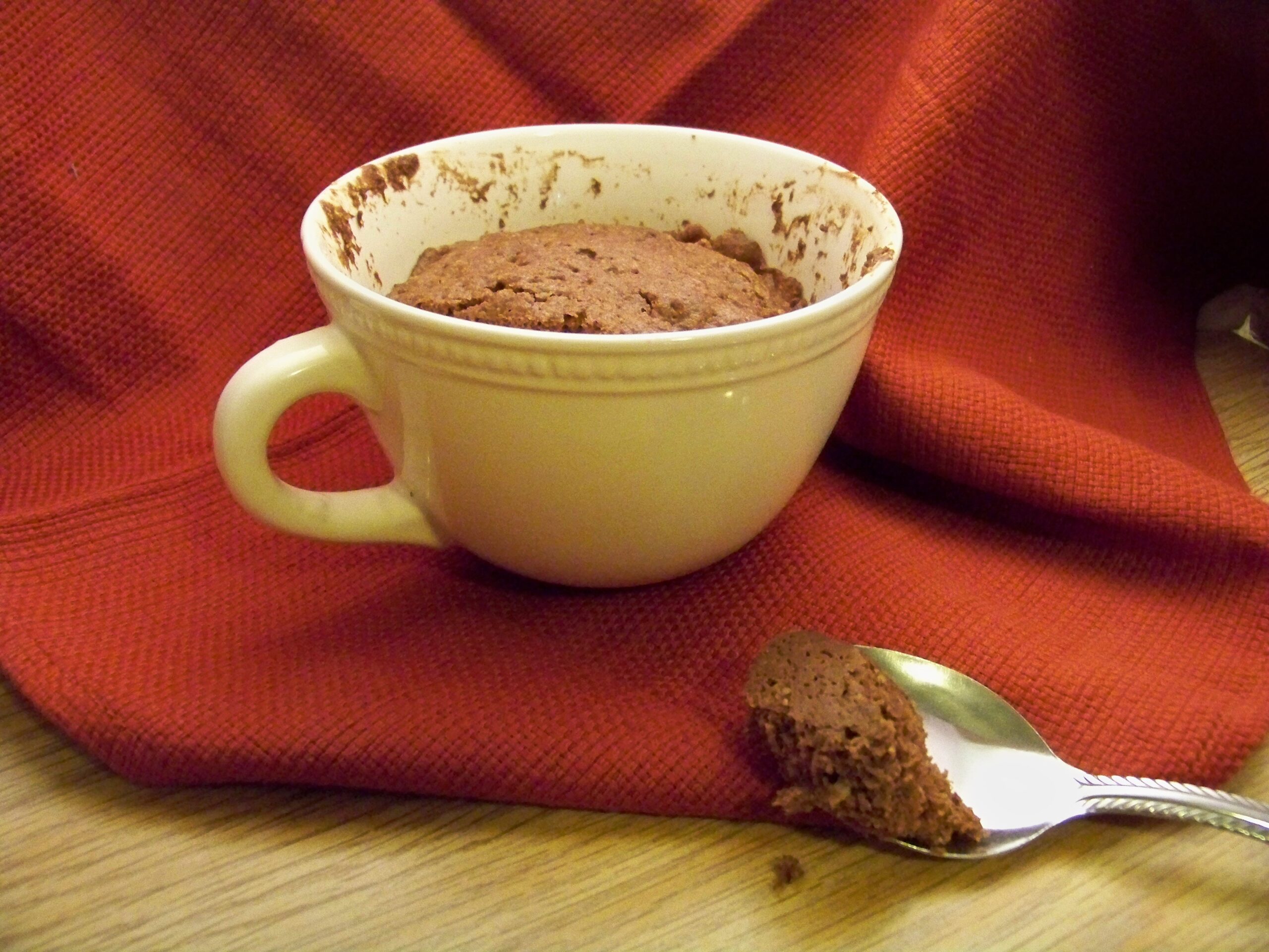 An image of a red cloth with a short white tea-cup style mug resting on it, containing a mocha muffin and a metal spoon slightly tot the right of the front of the mug with a ready bite of mocha muffin on it
