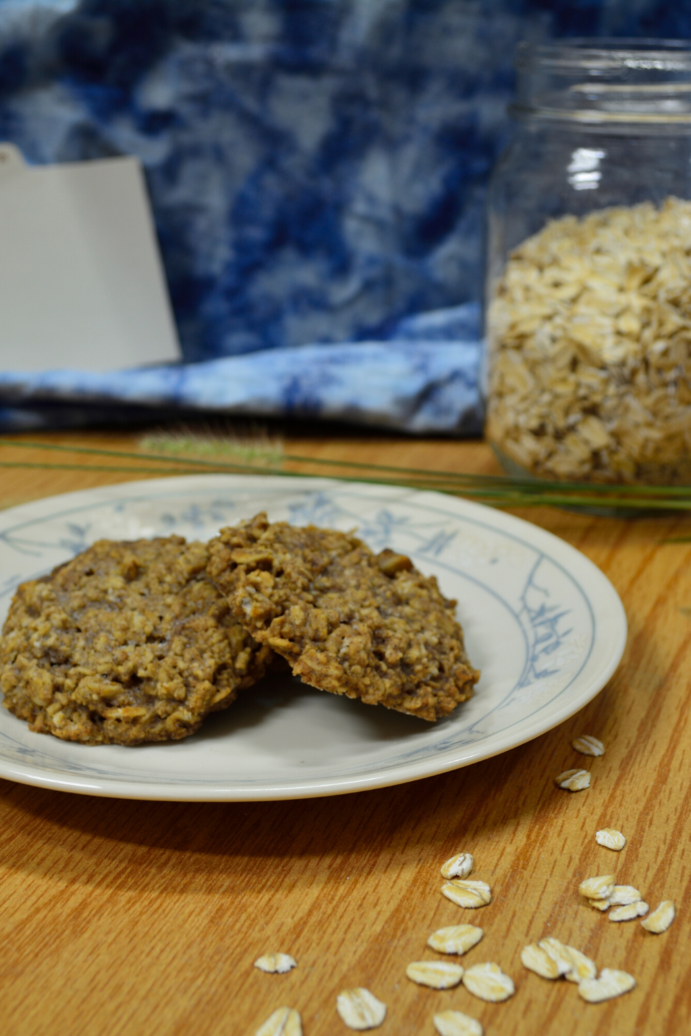Basic Oatmeal Cookies on a white plate with a wooden surface underneath, a cloudy looking blue cloth behind with a jar of oats and a recipe card behind