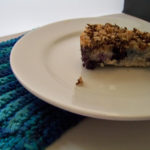 Plated blueberry buckle dessert on white plate with a potholder in shades of blue underneath the left side