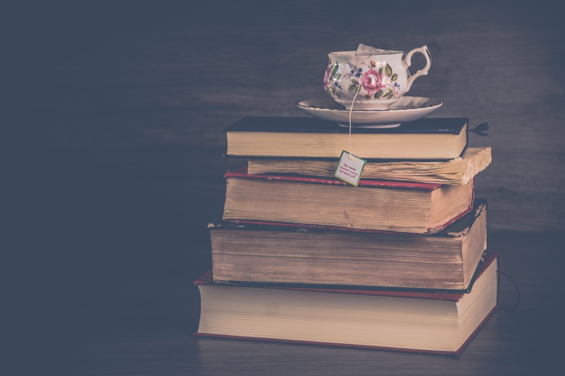 A dark background with a stack of four worn looking books, one quite tattered - page side facing us - with a china mug decorated with pink flowers on a saucer on top, a tea bag tag hanging down from the mug over the top of the stack of books