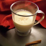 Sugar Free eggnog in a clear glass mug with a handle and a cinnamon stick in the front, red slightly glittery cloth in the background