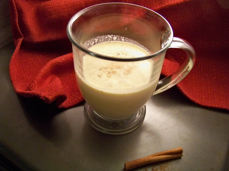 Sugar Free eggnog in a clear glass mug with a handle and a cinnamon stick in the front, red slightly glittery cloth in the background