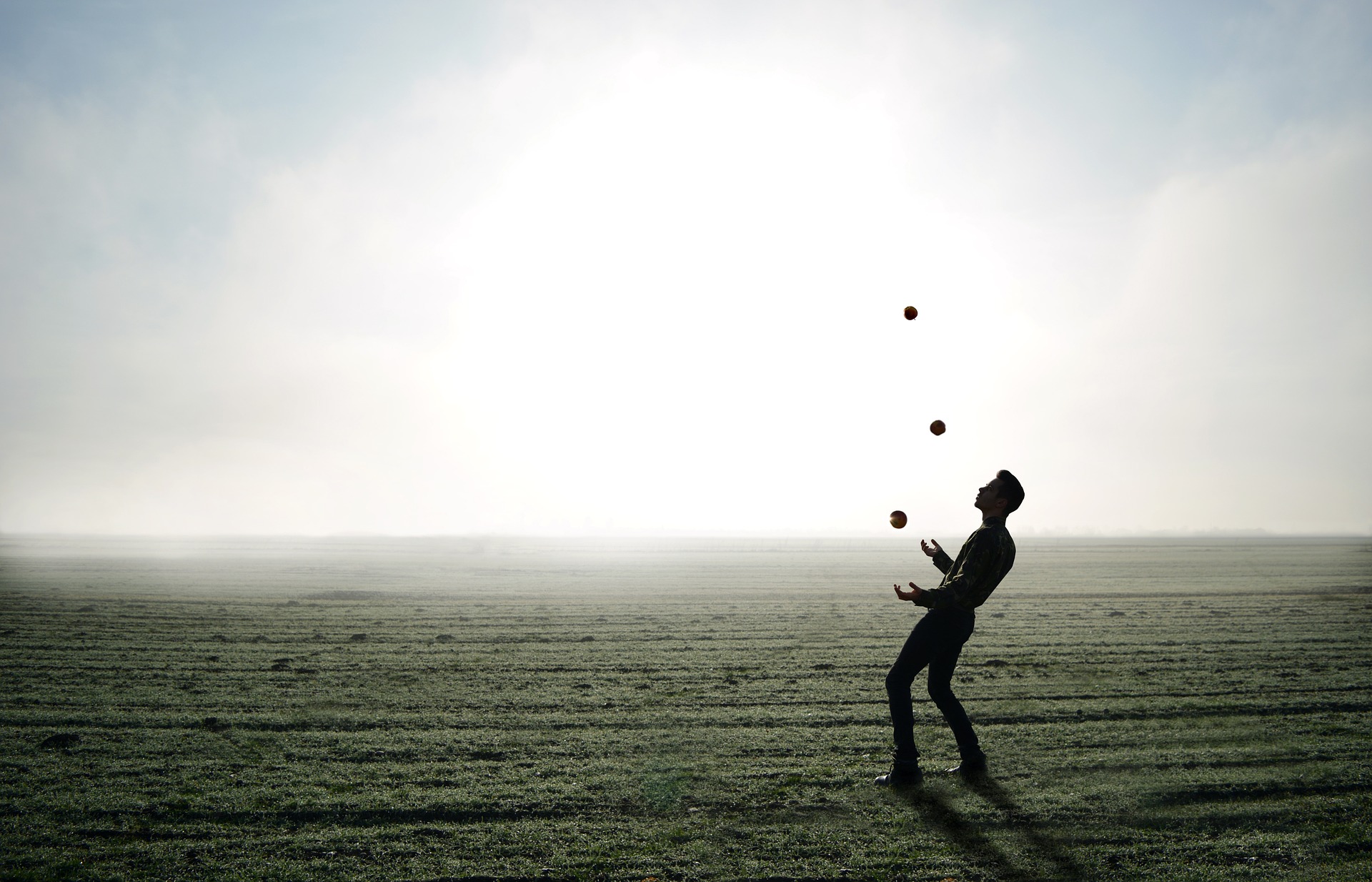 Landscape scene: a low mown field with the sky blending into it... a man leaning backwards and juggling three cylindrical objects standing in the field