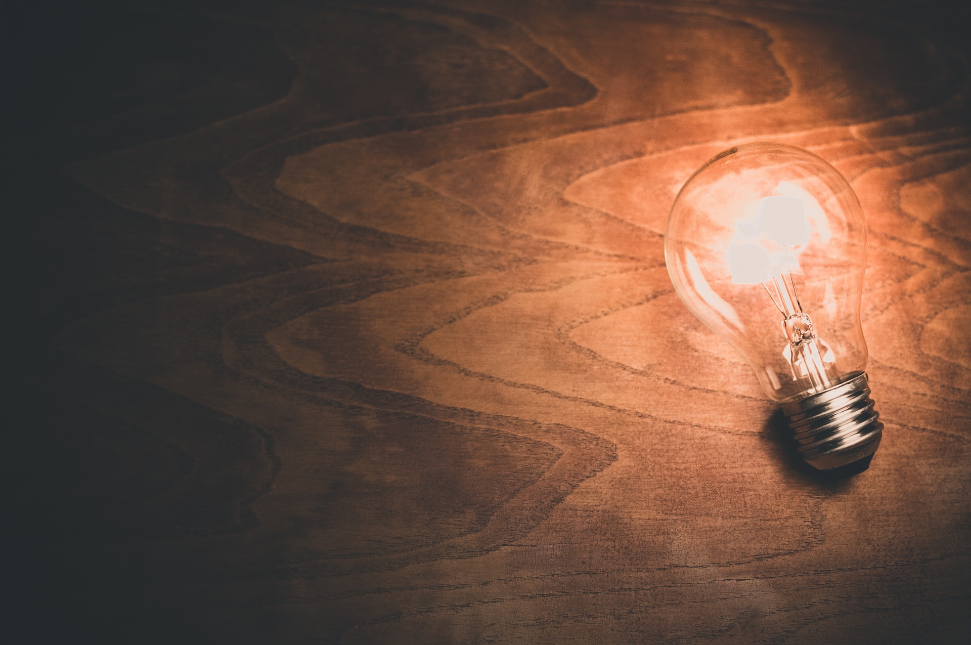 A wooden surface with a lit light bulb lying alone on it, illuminating the closest portion of the surface while the further portion of the surface remains dark