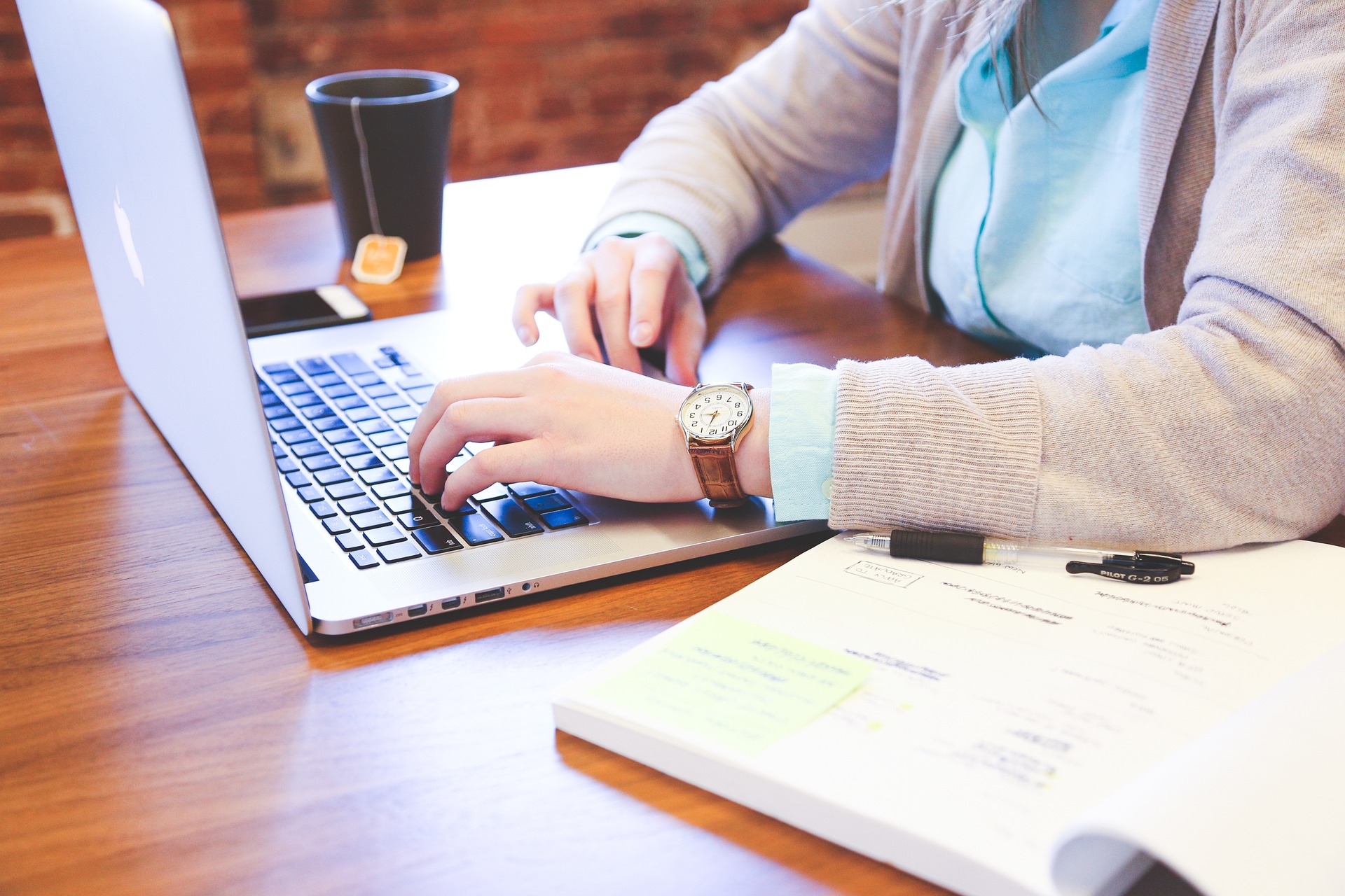 A figure with long sleeves and a brown strapped watch on the left wrist working on a grey laptop with a mug of tea, tag hanging from it in the background