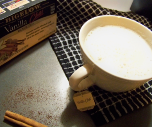 Vanilla Chai Tea Latte in a short white teacup with a tea tag hanging from the mug and a box of vanilla chai tea in the top left, a black and white checkered cloth under the mug and a cinnamon stick slightly visible in the bottom left