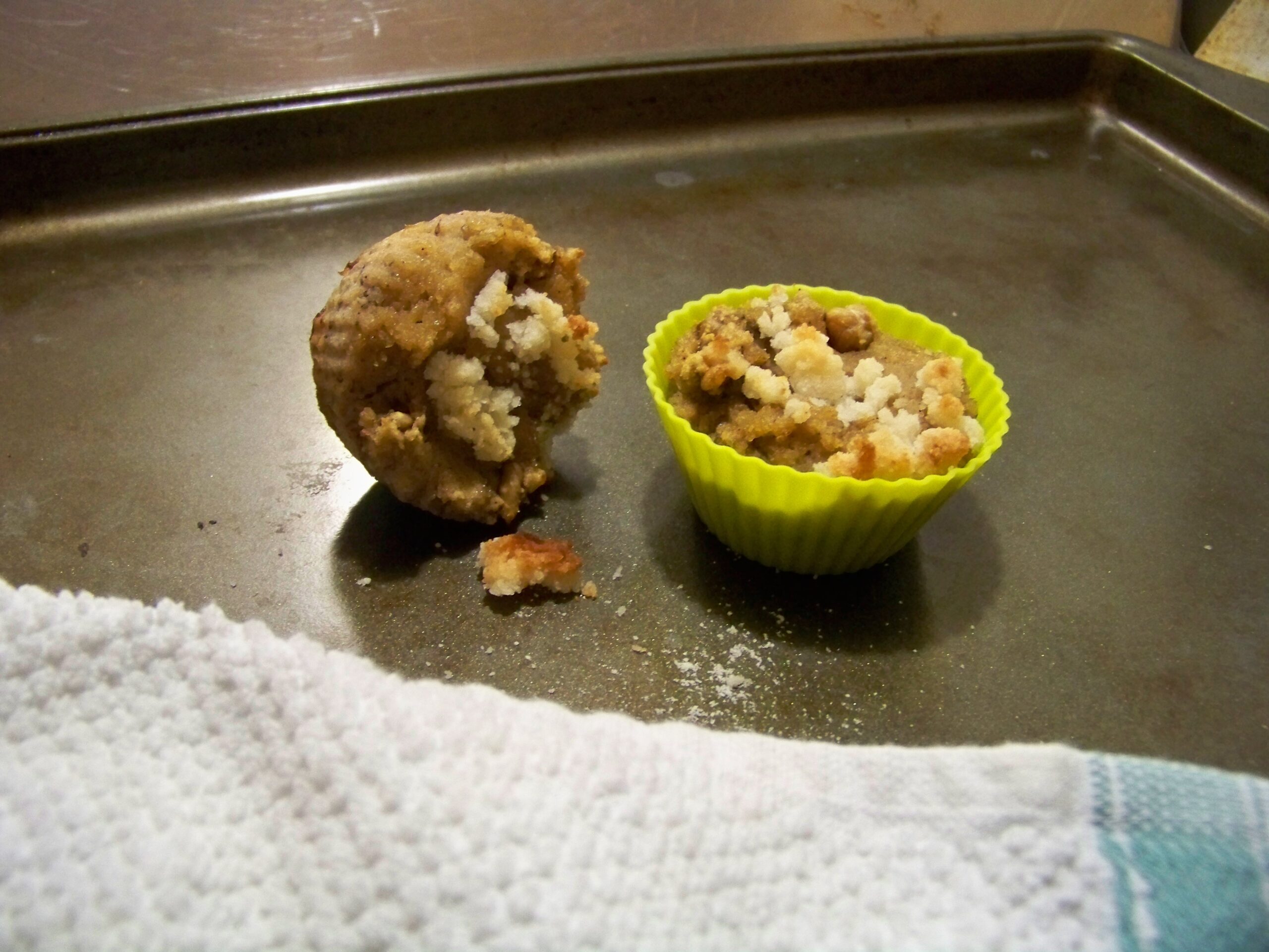 Two banana nut muffins, one in a yellow silicone liner and the other without a liner on a cookie sheet with a white cloth with a blue stripe in the forefront of the photo