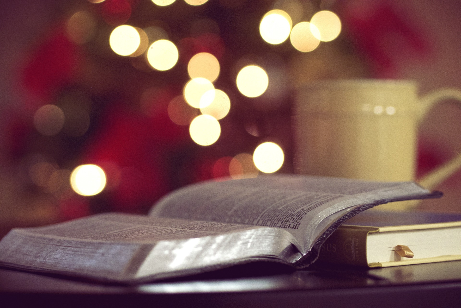 a blurry background with an open Bible in the forefront and a closed book underneath one side of the cover with a blurry yellow mug on the back right side of the image