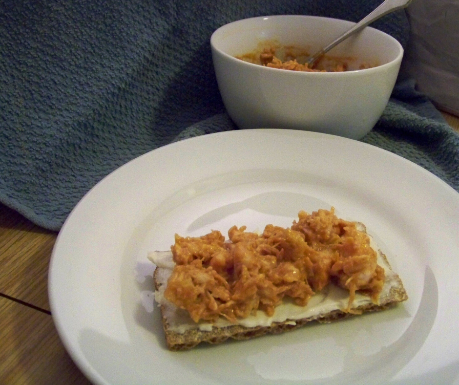 Tangy BBQ Chicken Salad on a wasa cracker on a white plate with a white bowl of extra chicken salad, a spoon coming from it on a blue cloth