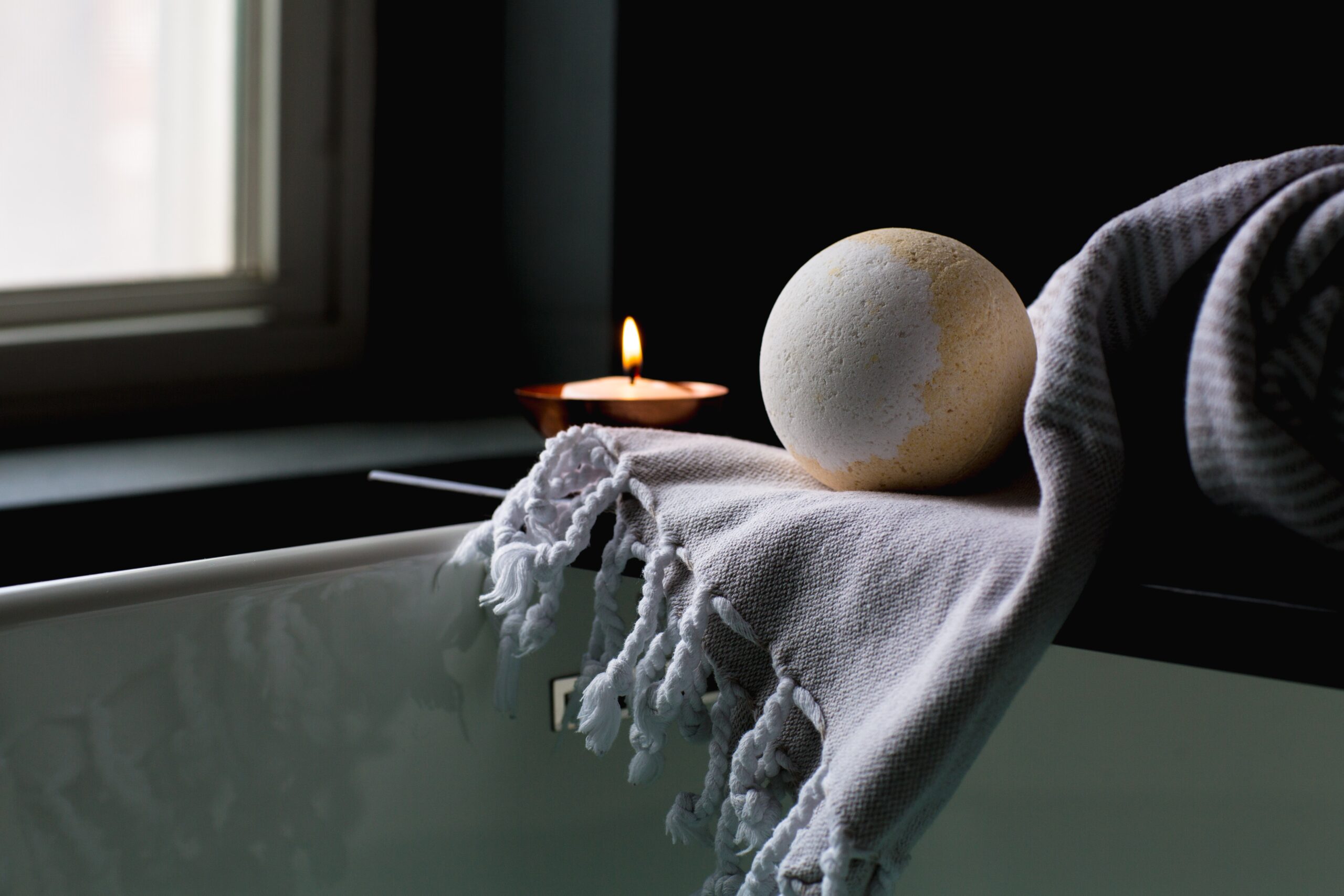 A lit tea light in a small dish with a orangish white bath bomb on a towel in the forefront