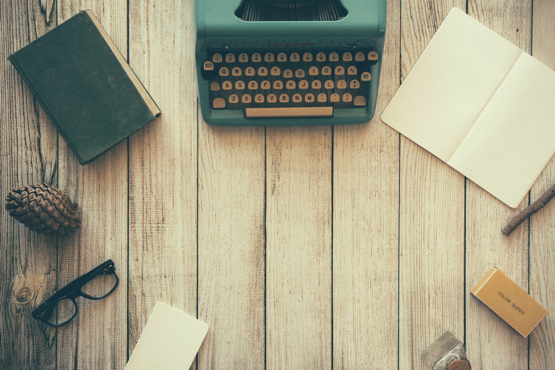 Wooden planks underneath an old fashioned blueish typewriter, an open blank notebook, a closed book, a pinecone, glasses, and some miscellaneous items