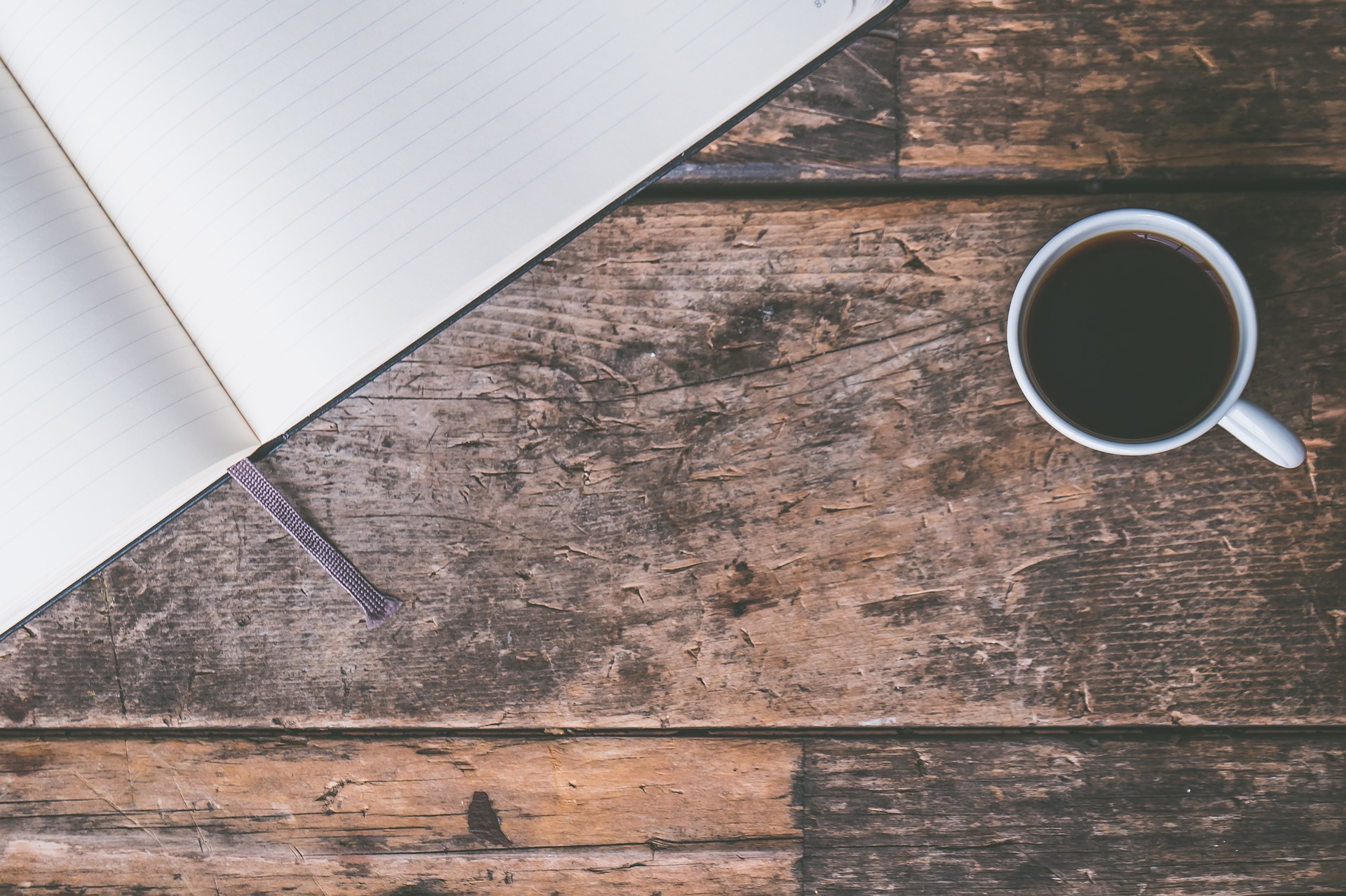 A dark wooden surface with an open lined notebook in the top left corner and a mug of black coffee towards the right