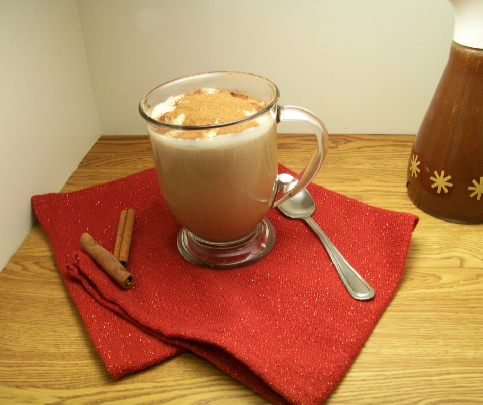 Gingerbread Coffee Syrup in an old fashioned syrup dispenser with a mug of gingerbread coffee in a clear mug with a spoon on one side and cinnamon sticks on the other, a red cloth underneath