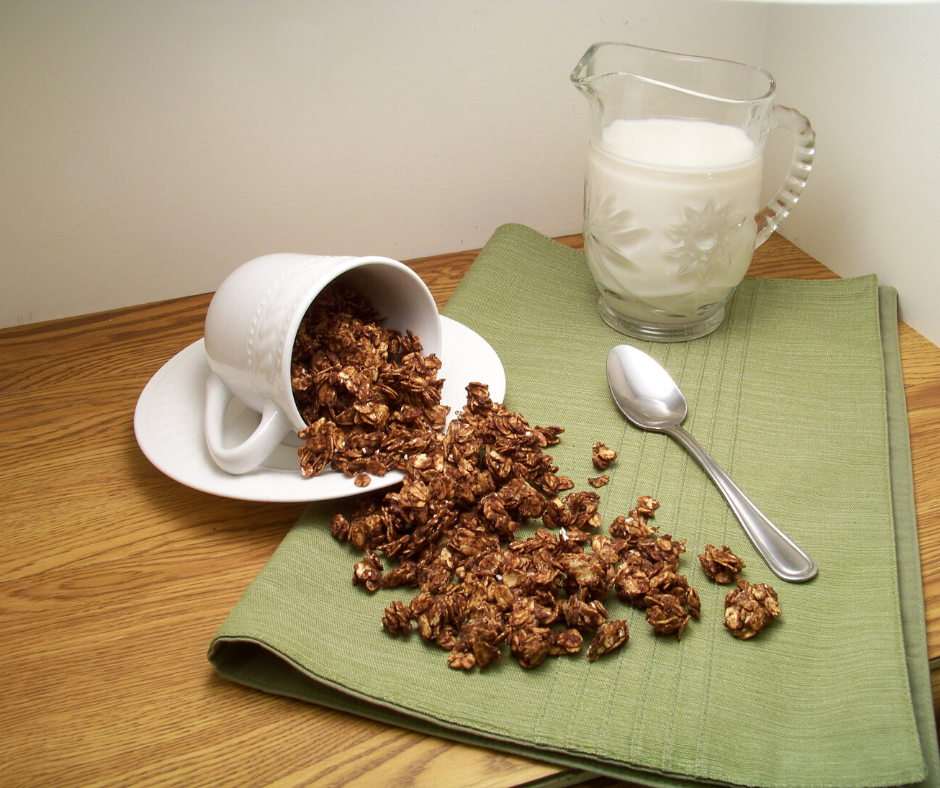 Peppermint Mocha Granola spilling from a white mug on a saucer laid sideways with a spoon beside the spillage, all on a green cloth with a small container of milk in a clear glass container with a handle and a spout in the back right