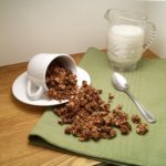 Peppermint Mocha Granola spilling from a white mug on a saucer laid sideways with a spoon beside the spillage, all on a green cloth with a small container of milk in a clear glass container with a handle and a spout in the back right