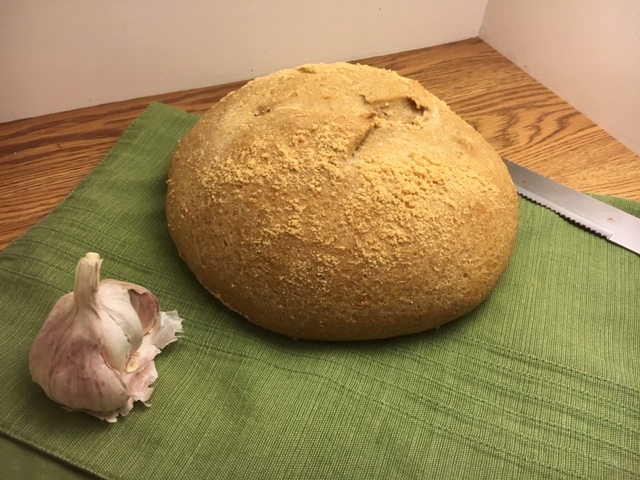 Garlic Parmesan Sourdough Bread whole and uncut, sitting on a green placemat with a bread knife beside it and a partial bulb of garlic to the front