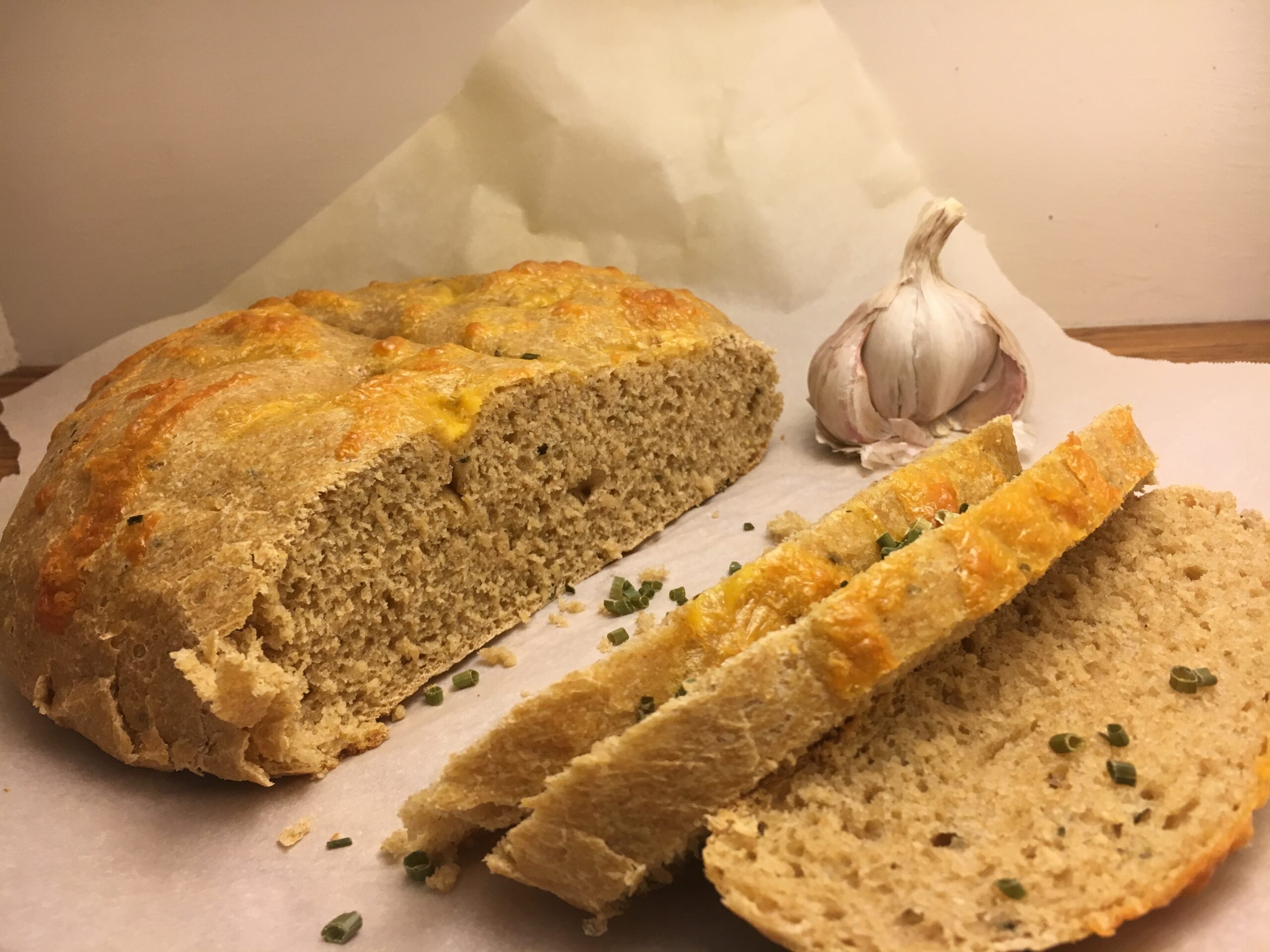 Garlic Cheddar Chive Sourdough Bread sliced on parchment paper with a bulb of garlic slightly behind