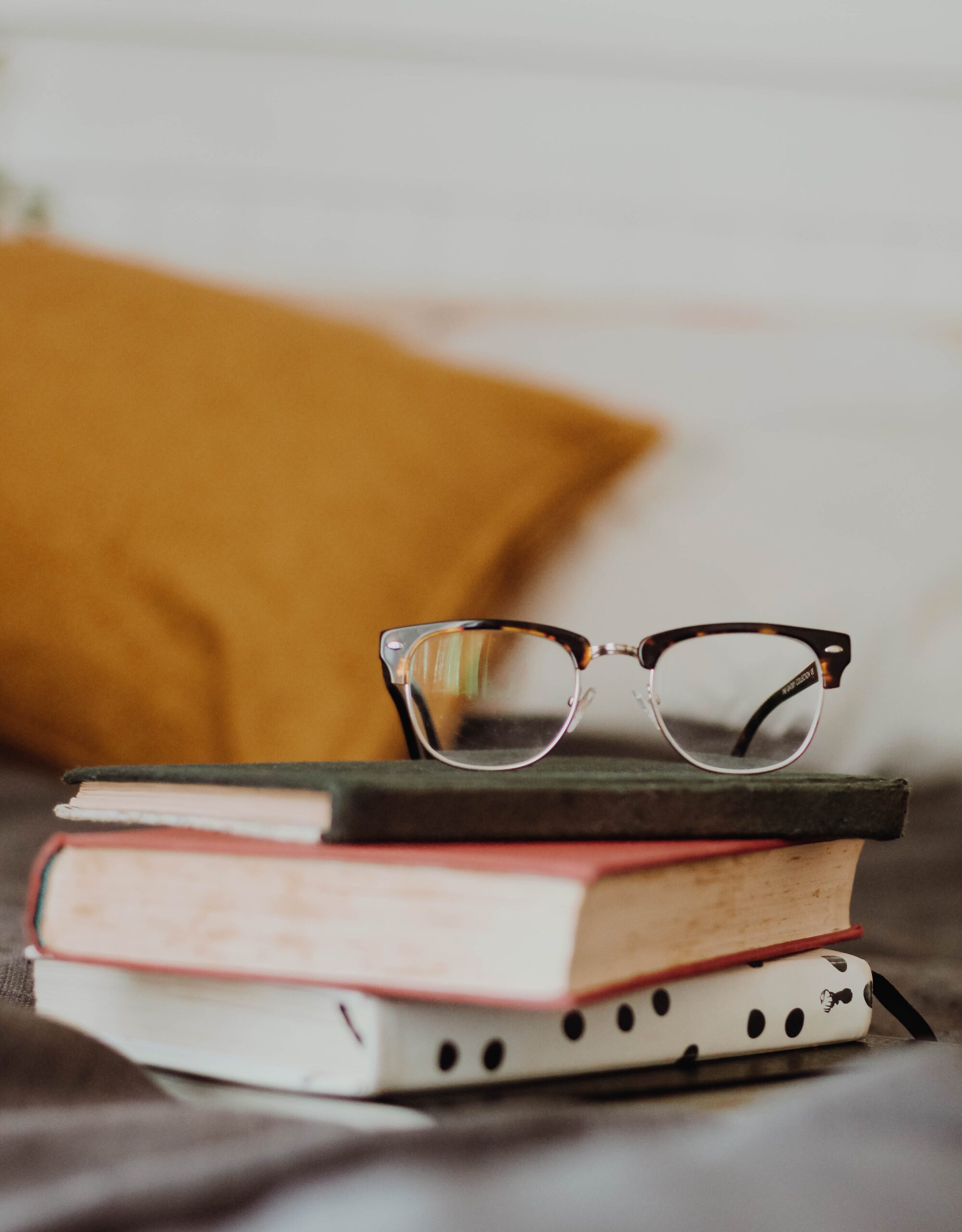 A small stack of books with glasses resting on top, an orange-ish pillow in the background