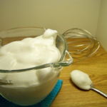 Whipped "Cream" in a clear bowl with a spoon of the "cream" beside it and a whisk in the background