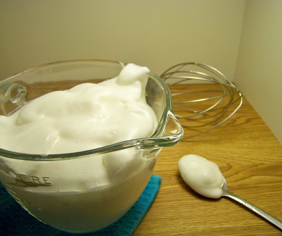 Whipped "Cream" in a clear bowl with a spoon of the "cream" beside it and a whisk in the background