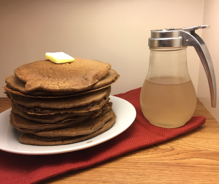 A stack of Dark Chocolate Sourdough Pancakes topped with a pat of butter on a round white plate on top of a red cloth with a syrup dispenser beside it