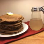 A stack of Dark Chocolate Sourdough Pancakes topped with a pat of butter on a round white plate on top of a red cloth with a syrup dispenser beside it