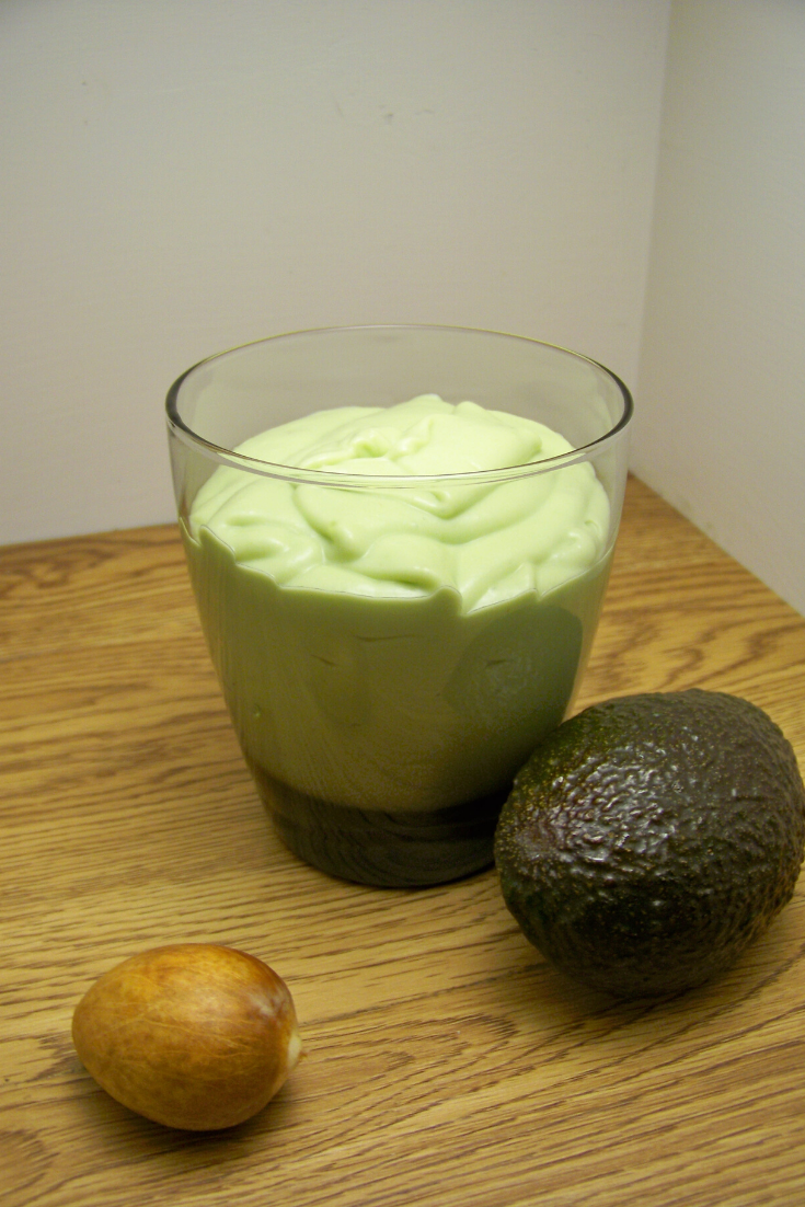 3 Ingredient Avocado Smoothie in a short juice glass on a wooden surface, an avocado beside the glass and an avocado pit to the front left of the glass
