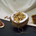 White Christmas Dreaming Granola in a opened coconut as a bowl with a wooden spoon holding hazelnuts to the left and a small dish holding whole coffee beans to the right
