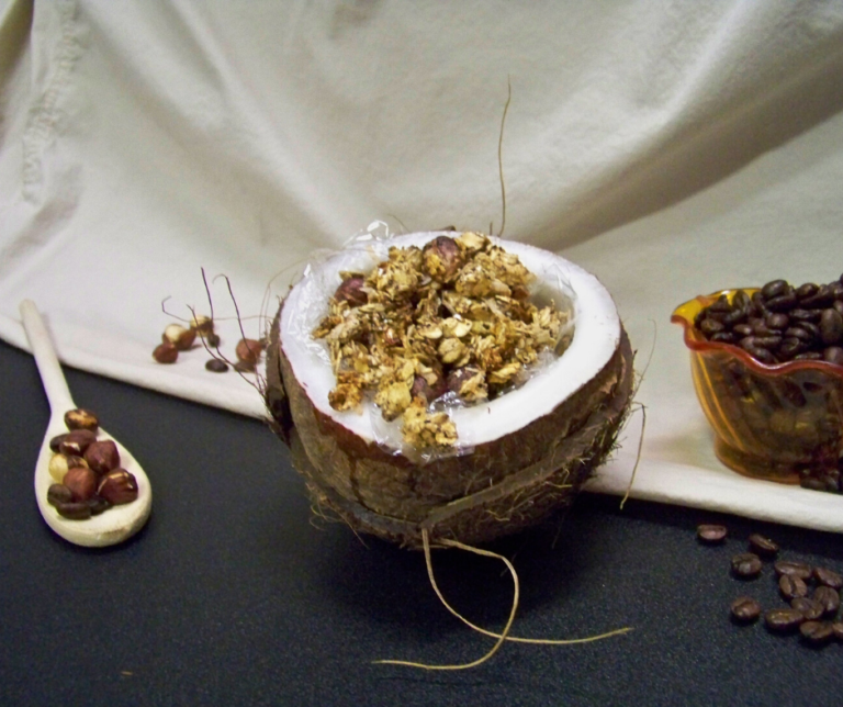 White Christmas Dreaming Granola in a opened coconut as a bowl with a wooden spoon holding hazelnuts to the left and a small dish holding whole coffee beans to the right