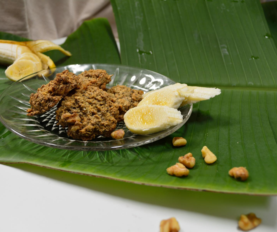 Banana Nut Bread Cookies plated on a clear plate with a banana leaf underneath, walnuts around and sliced banana in the background and on the plate