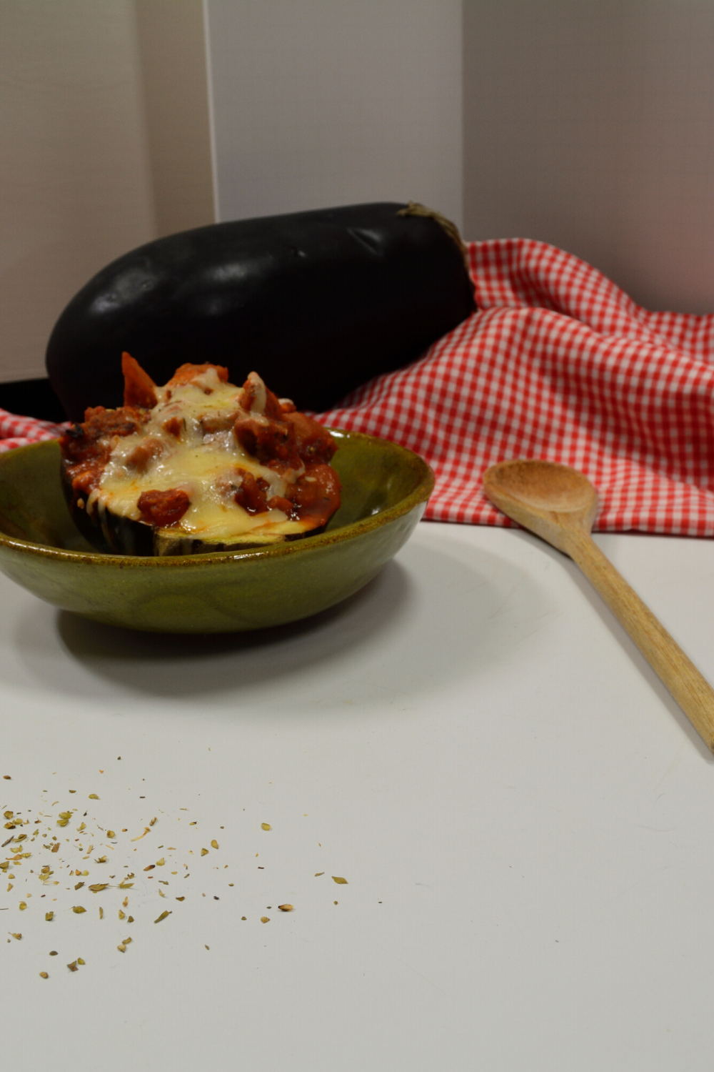Stuffed Eggplant in a homemade green ceramic bowl with a wooden spoon to the right and a checkered red and white cloth behind with a whole eggplant on top