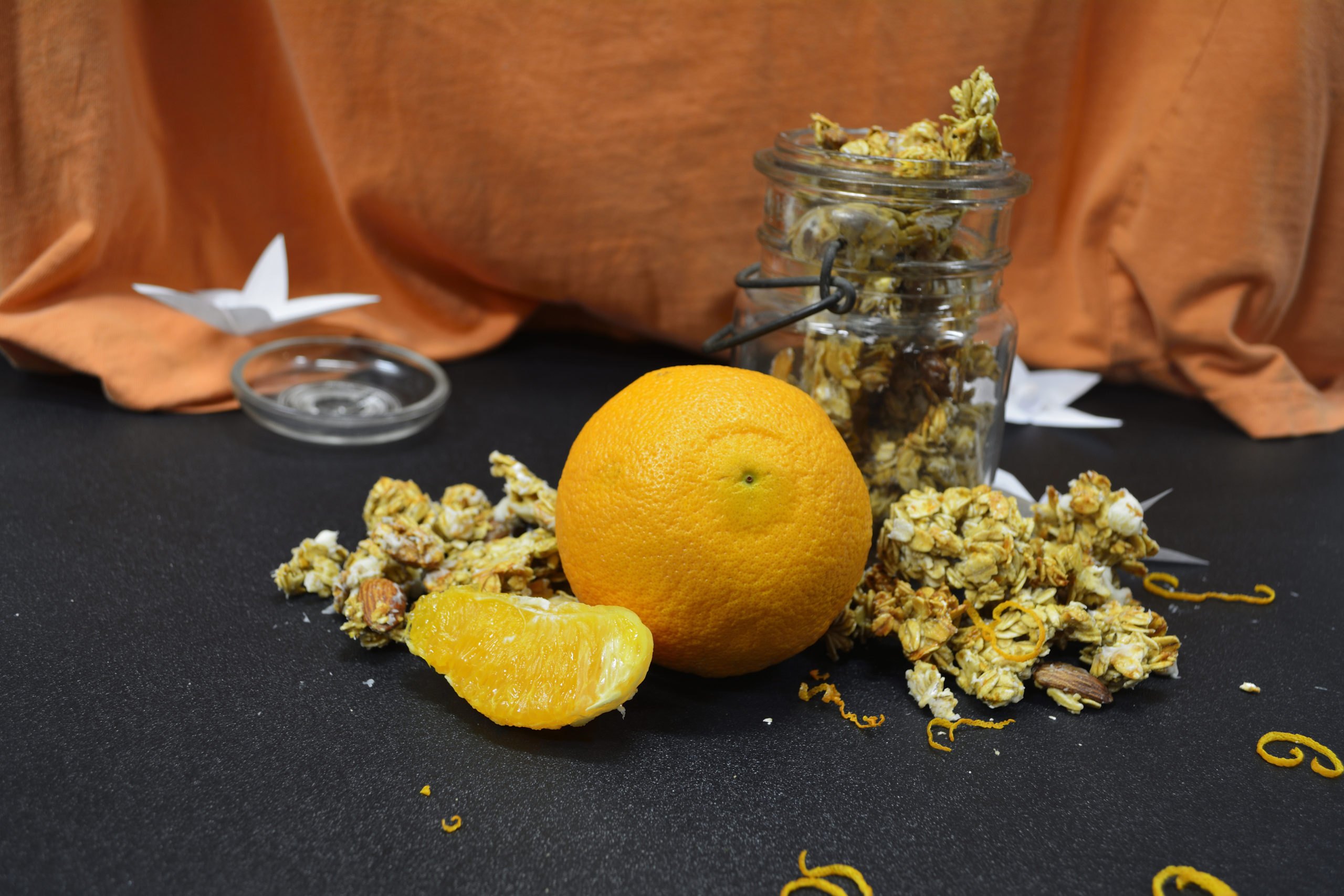 Orange Vanilla Granola spilling out of an antique mason jar, an orange fruit in the front and an orange cloth in the background
