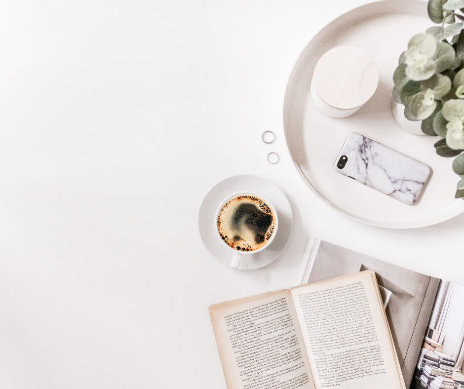 A white background with a white mug of black coffee on a white saucer, an open book and a round tray with a marble phone case and a plant slightly visible