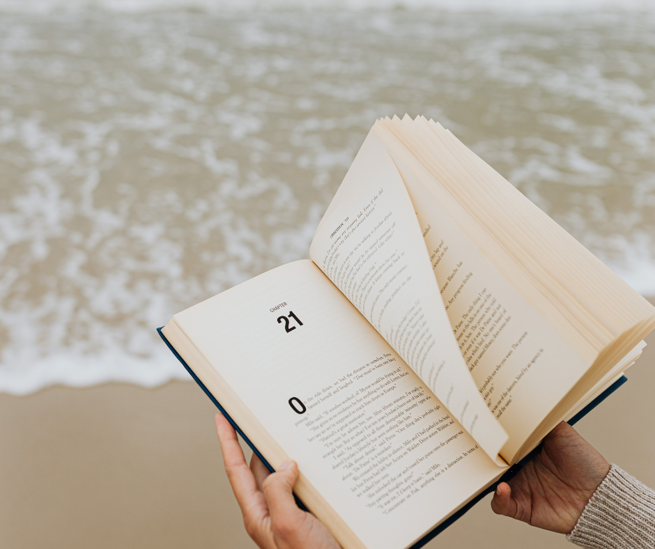 A person holding an open book by the water of a beach