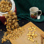 Apple Mulling Granola spilling from a cookie jar lid askew on the jar with an apple mug with a tea bag coming from it to the right and a wooden board with dehydrated apple pieces and cinnamon sticks to the front