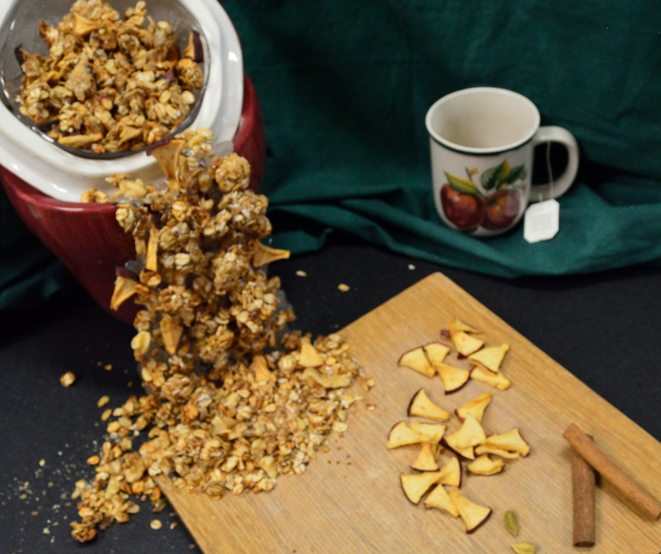 Apple Mulling Granola spilling from a cookie jar lid askew on the jar with an apple mug with a tea bag coming from it to the right and a wooden board with dehydrated apple pieces and cinnamon sticks to the front