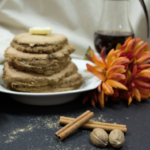 Oat Bran Gingerbread Pancakes stacked on a white plate with a pat of butter on top, syrup in a dispenser in the background with an orange flower to the front and whole nutmeg and cinnamon sticks at the forefront of the image