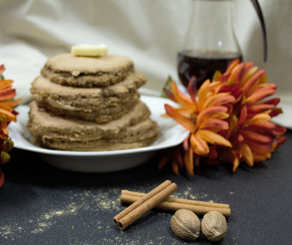 Oat Bran Gingerbread Pancakes stacked on a white plate with a pat of butter on top, syrup in a dispenser in the background with an orange flower to the front and whole nutmeg and cinnamon sticks at the forefront of the image