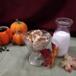 decorative pumpkins, one mug style containing coffee and coffee beans scattered in front, a glass ice cream dish with pumpkin latte ice cream and gingersnap crumbs beside an antique cream container full of milk with fall leaves in front on a green placemat with a burgundy background