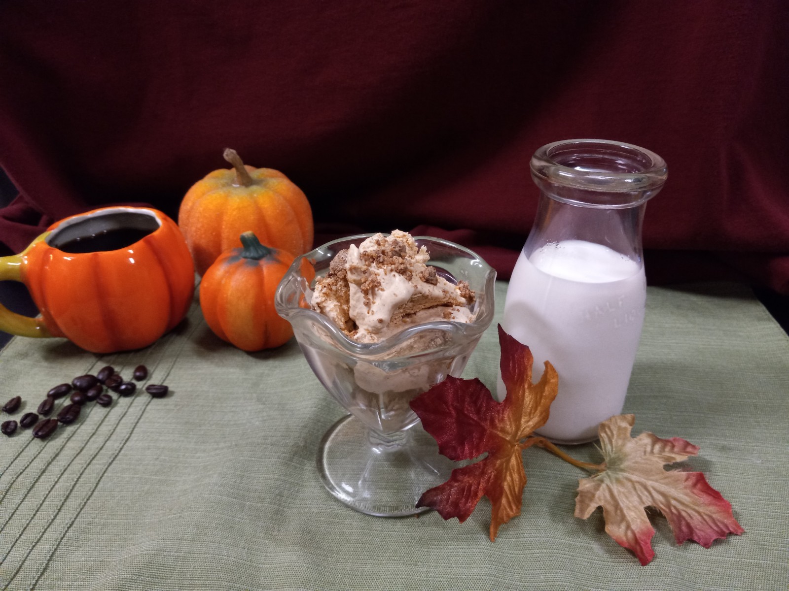 decorative pumpkins, one mug style containing coffee and coffee beans scattered in front, a glass ice cream dish with pumpkin latte ice cream and gingersnap crumbs beside an antique cream container full of milk with fall leaves in front on a green placemat with a burgundy background