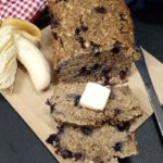 A sliced loaf of blueberry banana oat bread with a peeled banana on one side and a butter knife on the other on a black surface with a wooded cutting board under the bread