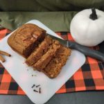 An orange and black checkered cloth under a white cutting board holding a sliced loaf of overnight soaked pumpkin bread with a knife beside it and a white pumpkin in front of the green cloth background