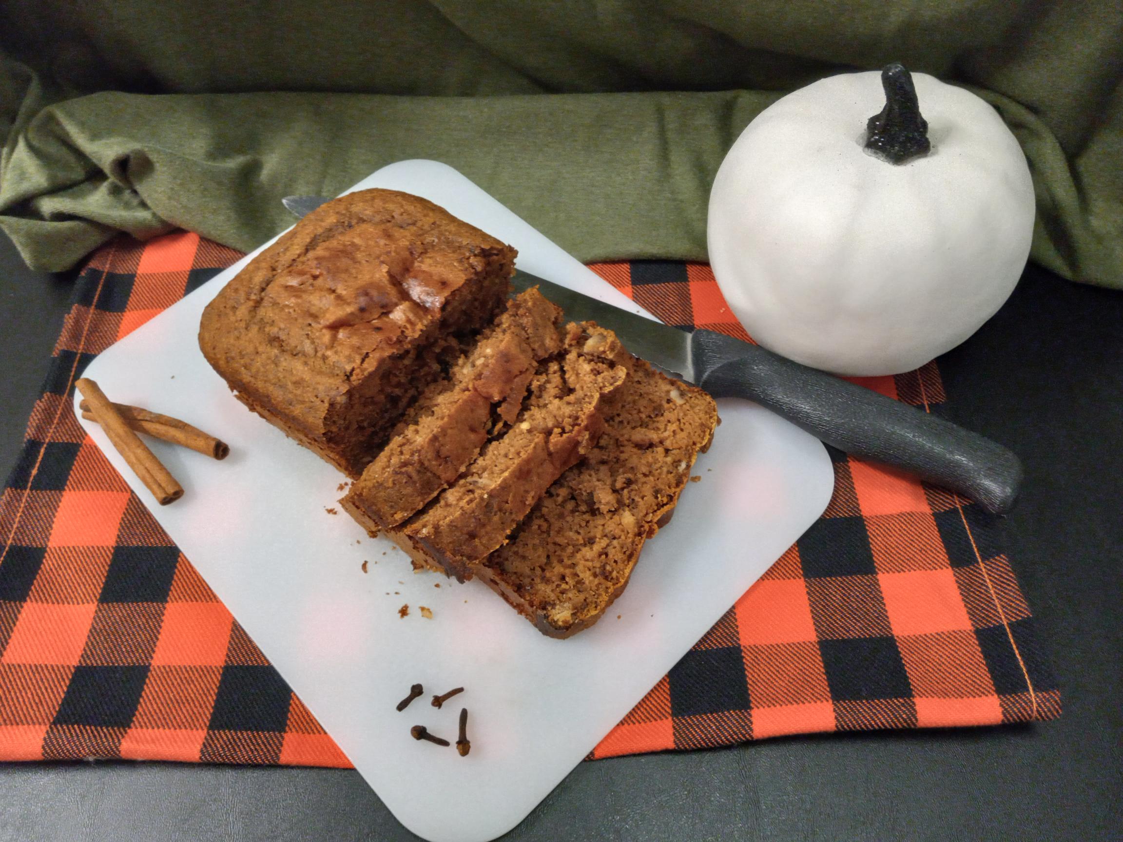 An orange and black checkered cloth under a white cutting board holding a sliced loaf of overnight soaked pumpkin bread with a knife beside it and a white pumpkin in front of the green cloth background