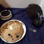 bluish purple surface with a plate of blueberry coconut pancakes, a bowl of fresh blueberries, and syrup in front of a coconut on it