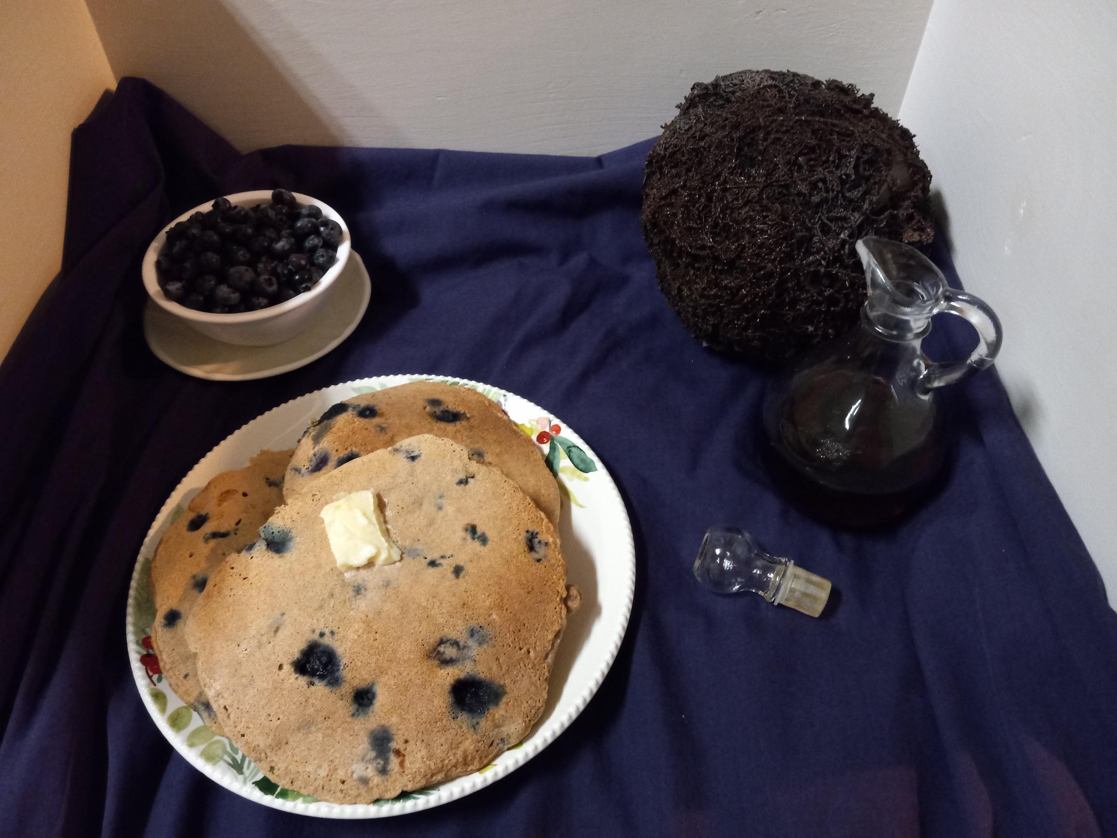 bluish purple surface with a plate of blueberry coconut pancakes, a bowl of fresh blueberries, and syrup in front of a coconut on it