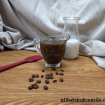 A red spatula on a wooden surface with a container of almond milk behind a small glass dish of coffee caramel sauce, coffee beans scattered in front