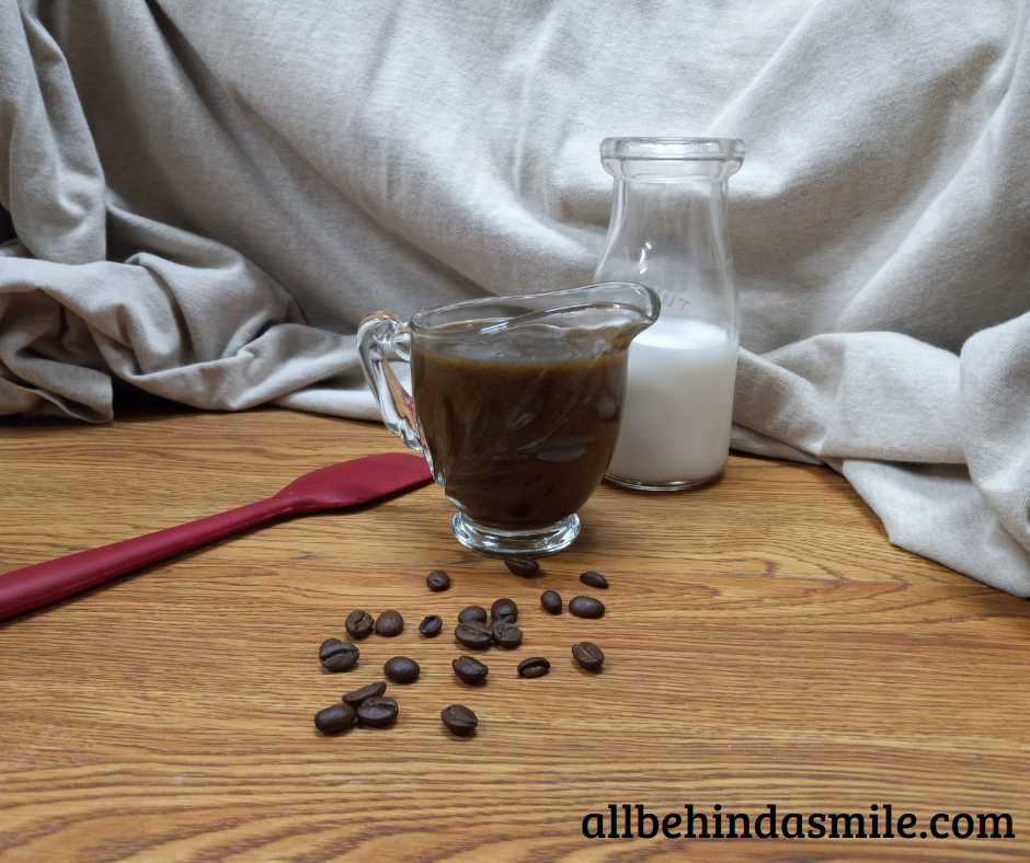 A red spatula on a wooden surface with a container of almond milk behind a small glass dish of coffee caramel sauce, coffee beans scattered in front